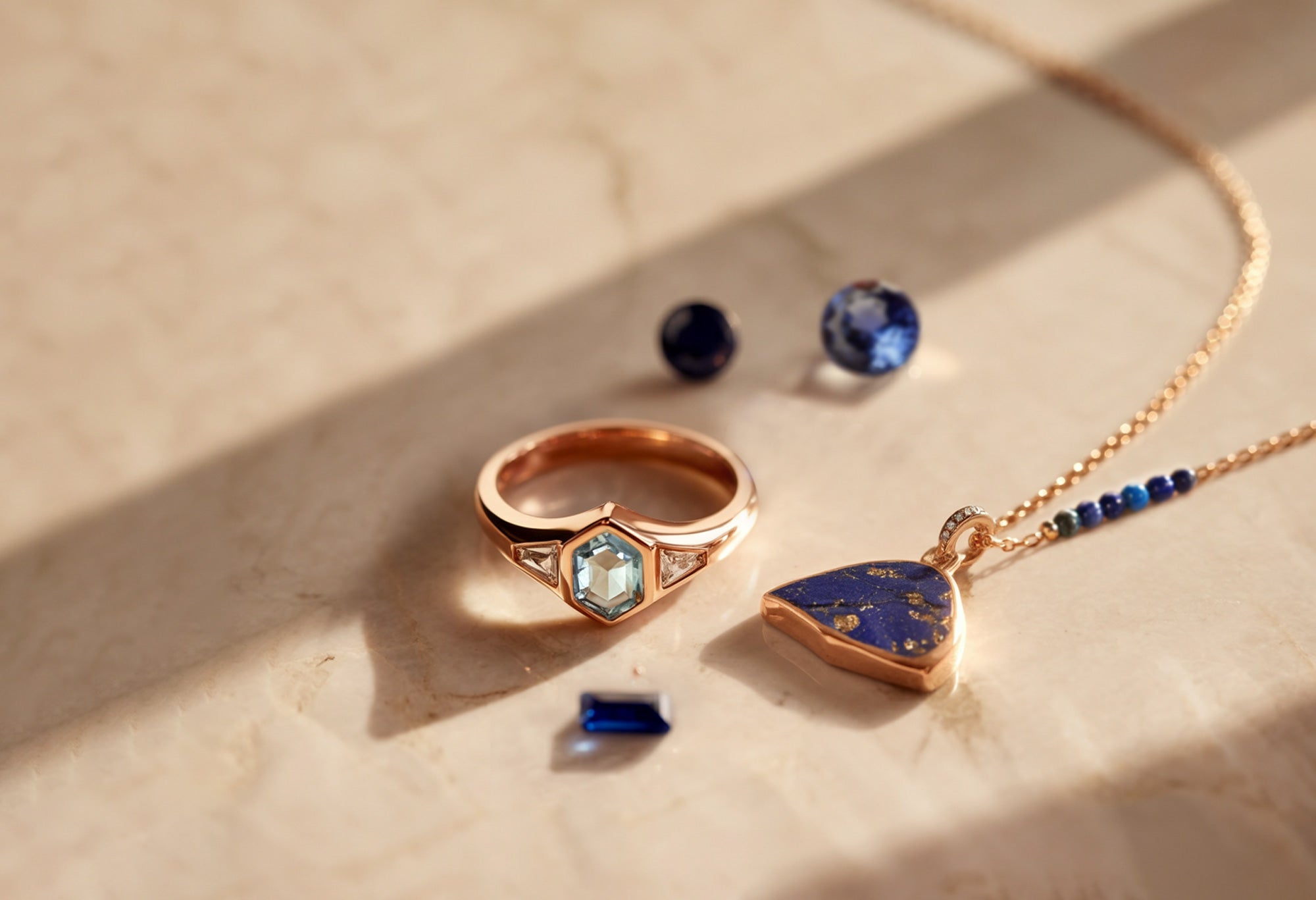 Chevalière en or rose sertie d’une aigue-marine et de deux diamants, accompagnée d’un pendentif en lapis lazulis et de pierres précieuses bleues non serties sur fond de marbre.