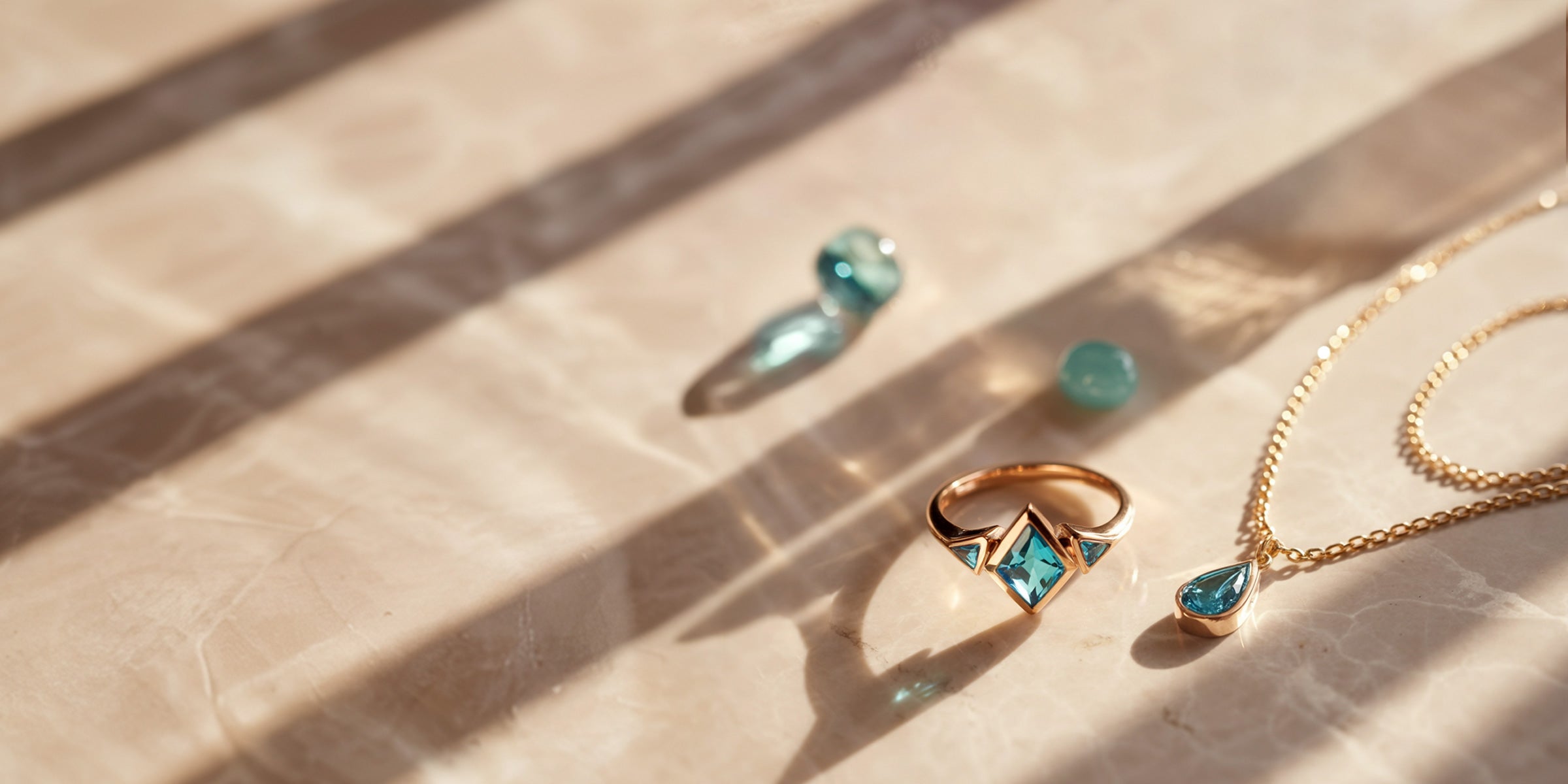 Photographie artistique de pierres précieuses turquoise comprenant une bague en or rose, un pendentif en tourmaline Paraiba, une amazonite et une aigue-marine sur fond de marbre avec jeux de lumière.