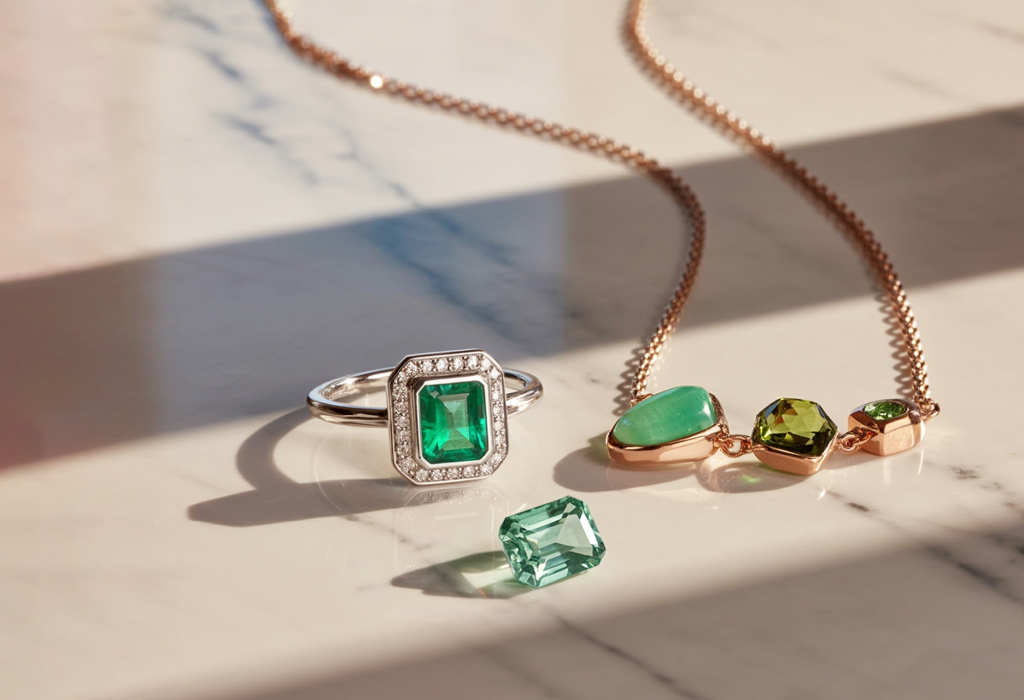 Bague en or blanc sertie d'une pierre précieuse verte émeraude et collier en or rose avec pierres vertes sur fond de marbre