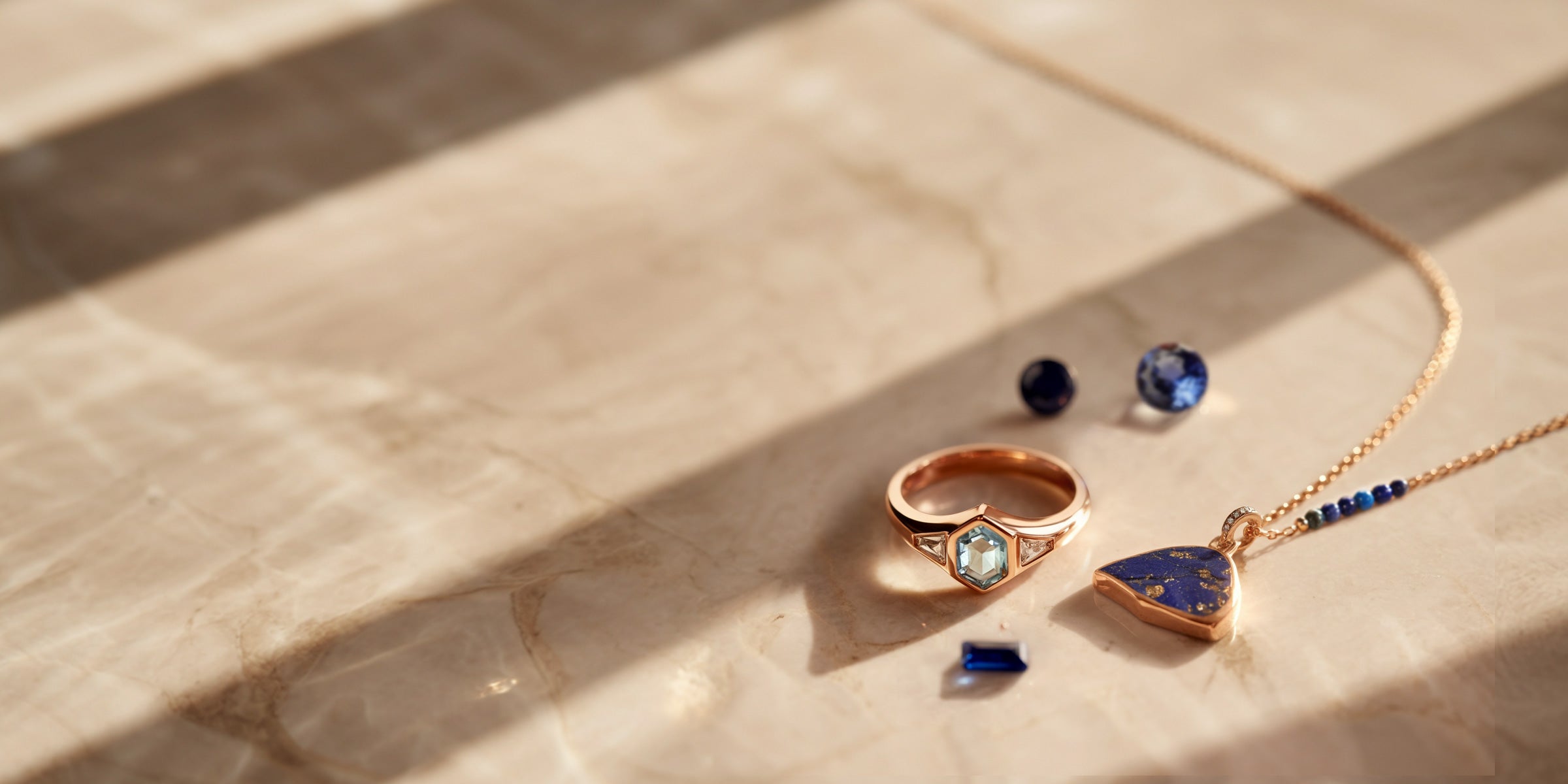 Bijoux en or rose avec pierres précieuses bleues : aigue-marine, tranche de lapis lazuli, spinelle bleu et saphir rond disposés sur une surface en marbre crème.